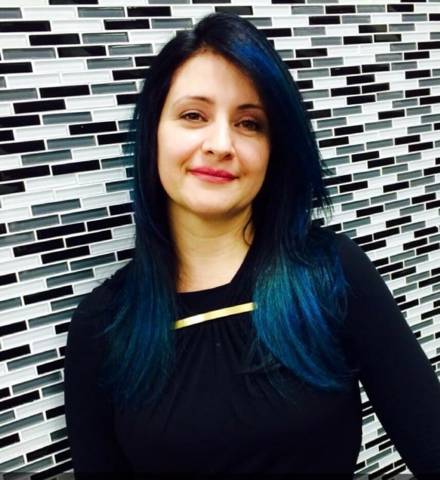 “Woman with long, straight black hair featuring vibrant blue highlights, wearing a black top, smiling in front of a tiled wall.”