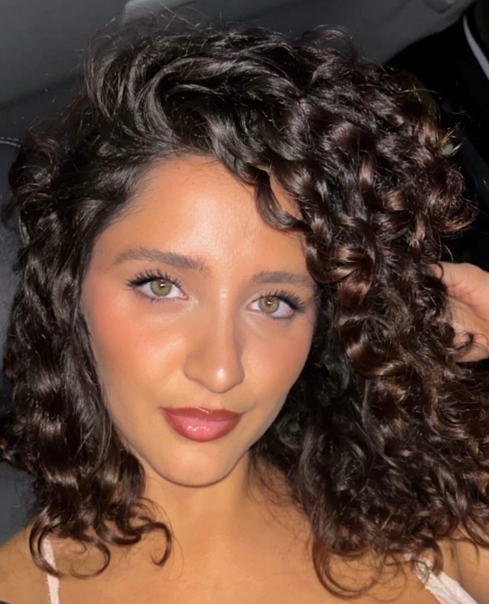 “Close-up of a woman with voluminous, curly dark hair, glowing skin, and natural makeup, smiling softly in a nighttime setting.”