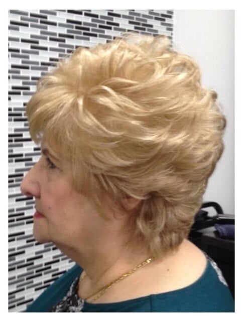 “Profile of a woman with a short, voluminous blonde hairstyle, styled in soft waves, photographed against a tiled background.”