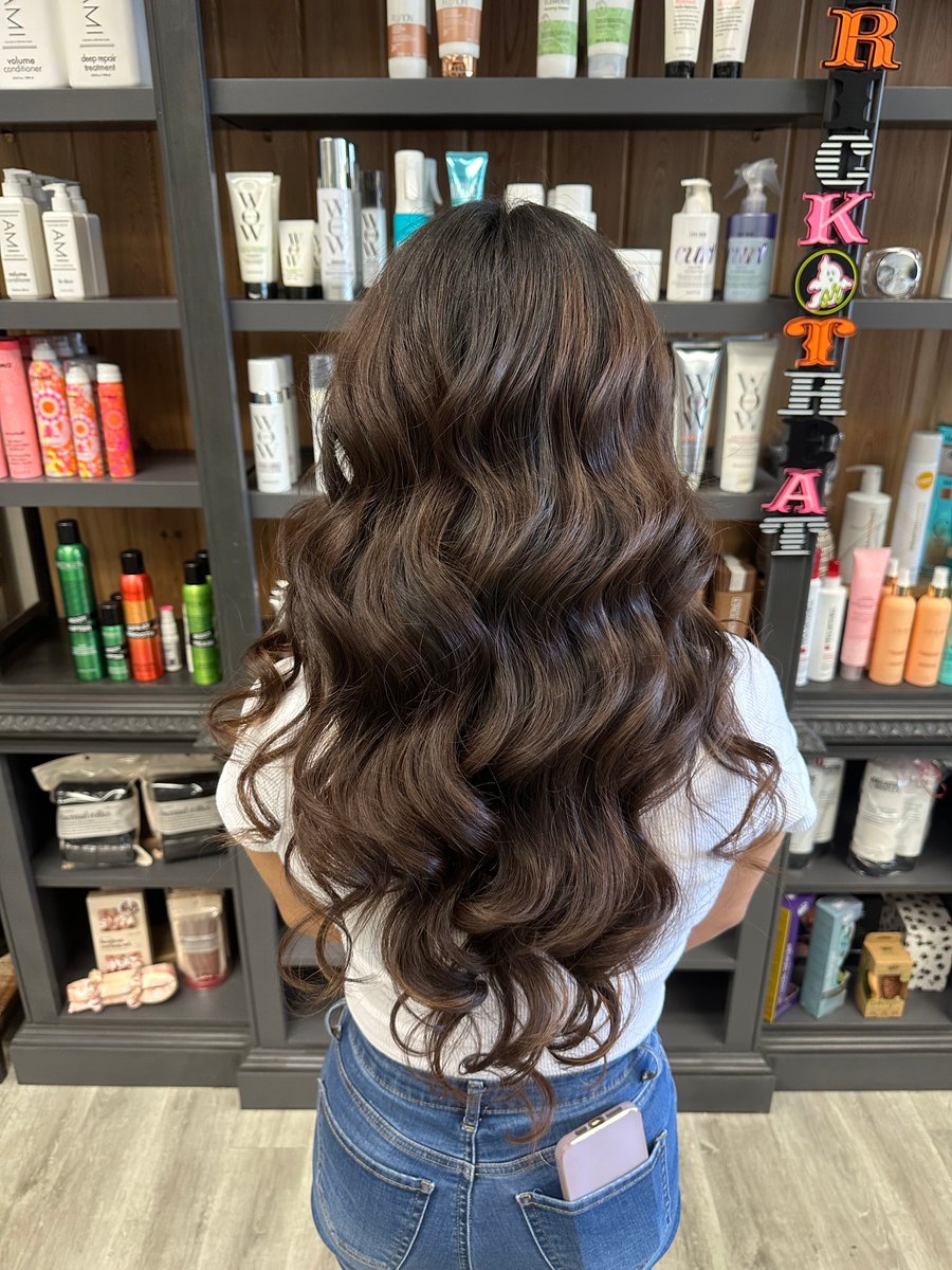 “Long, dark brown hair styled in voluminous curls, photographed in a salon with shelves of hair products in the background.”