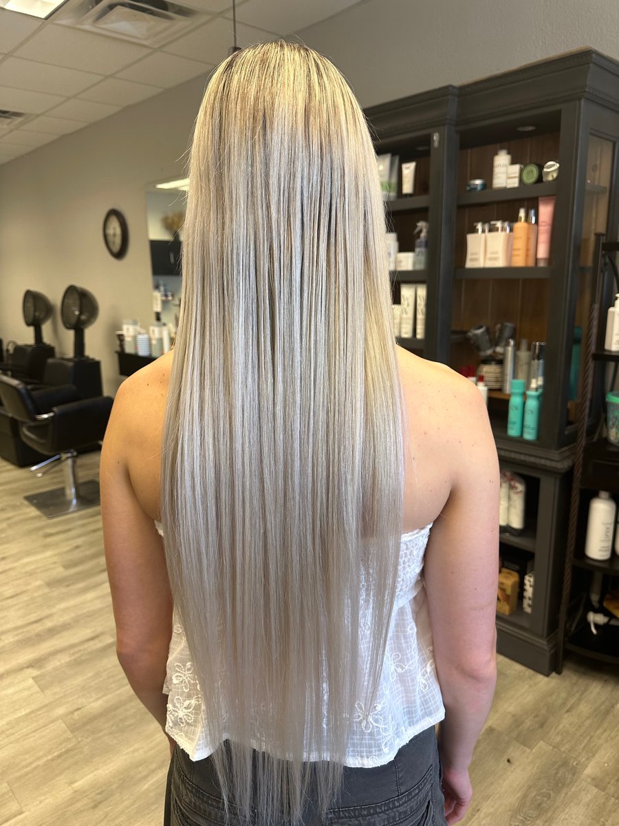 “Long, sleek, platinum blonde hair styled straight, shown from the back in a salon with shelves of products in the background.”