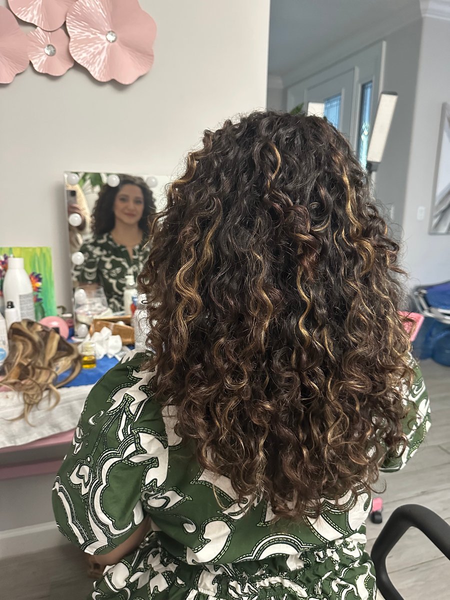 “Curly hair with subtle highlights styled naturally, shown from the back, with a reflection in a mirror and a vibrant salon setting.”